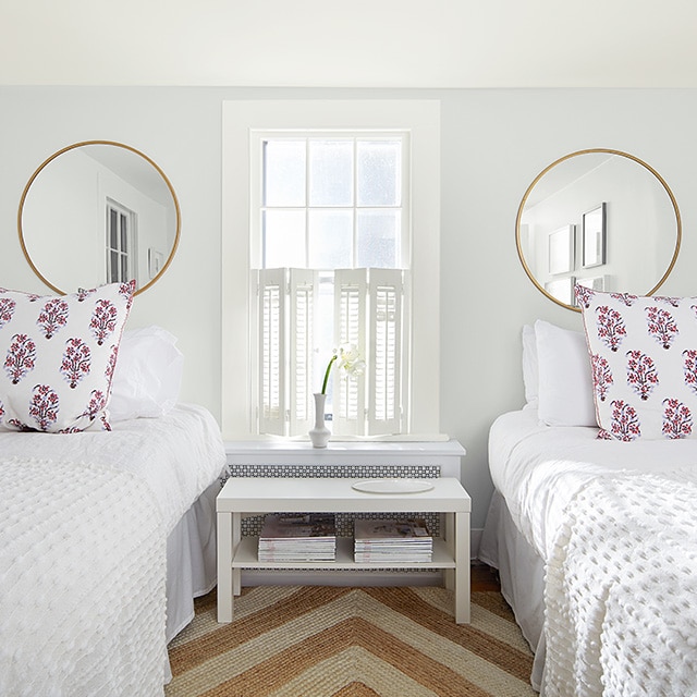Chambre à coucher blanche avec deux miroirs circulaires au mur, lits jumeaux à la literie blanche, fenêtre blanche avec persiennes et tapis à rayures en jute. Chambre à coucher blanche avec deux miroirs circulaires au mur, lits jumeaux à la literie blanche, fenêtre blanche avec persiennes et tapis à rayures en jute.