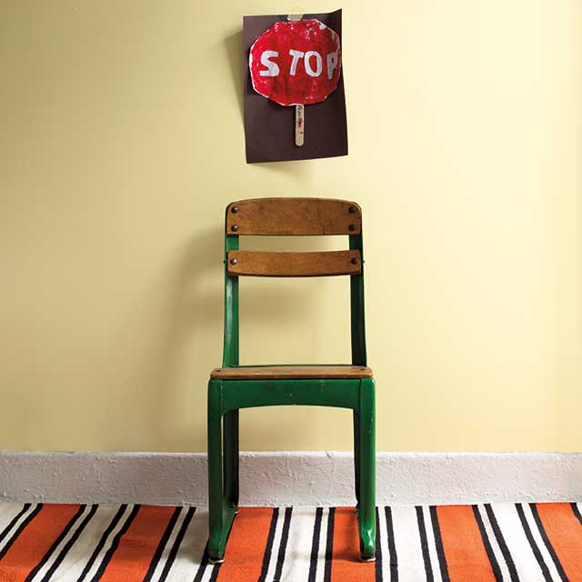 A green and wooden school chair against a soft yellow painted wall, with a hand-painted stop sign on the wall, and an orange, white, and black striped rug. A green and wooden school chair against a soft yellow painted wall, with a hand-painted stop sign on the wall, and an orange, white, and black striped rug.