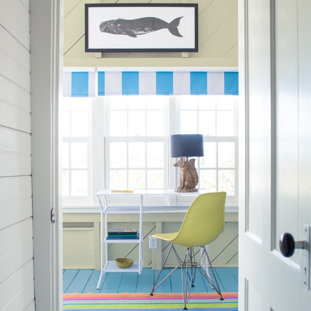 Couloir blanc avec porte blanche qui ouvre sur un bureau à l’ambiance de bord de mer avec mur et plafond en planches à feuillure jaune pâle, tableau de baleine suspendu au-dessus de plusieurs fenêtres, table blanche, chaise jaune et plancher bleu. Couloir blanc avec porte blanche qui ouvre sur un bureau à l’ambiance de bord de mer avec mur et plafond en planches à feuillure jaune pâle, tableau de baleine suspendu au-dessus de plusieurs fenêtres, table blanche, chaise jaune et plancher bleu.