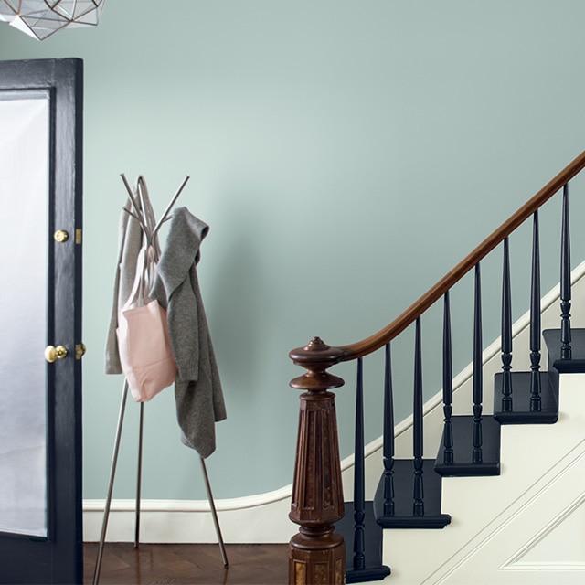A pretty, navy-blue painted entryway staircase with a wood handrail and newel post next to a bluish gray painted wall with white trim, and a coatrack at the bottom of the stairs. A pretty, navy-blue painted entryway staircase with a wood handrail and newel post next to a bluish gray painted wall with white trim, and a coatrack at the bottom of the stairs.