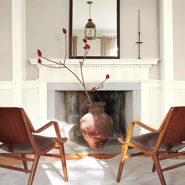 A serene living room with a greige-painted upper wall, white lower wall, mantel, and molding, a mirror, clay vase, and wood chairs on a white rug. A serene living room with a greige-painted upper wall, white lower wall, mantel, and molding, a mirror, clay vase, and wood chairs on a white rug.