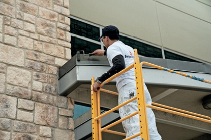 A Benjamin Moore painting contractor standing on yellow scaffolding and painting a facility exterior with Corotech® COMMAND®. A Benjamin Moore painting contractor standing on yellow scaffolding and painting a facility exterior with Corotech® COMMAND®.